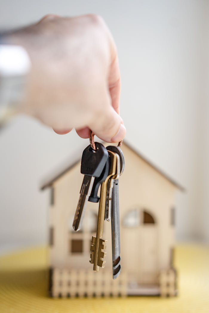 services-04 Close-up of a hand holding keys with a miniature wooden house in the background, symbolizing real estate investment.